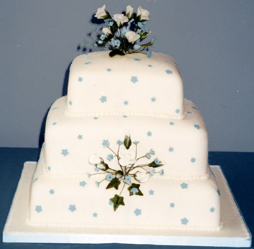 A three-tiered, stacked fruit and sponge wedding cake decorated with blue and ivory roses.